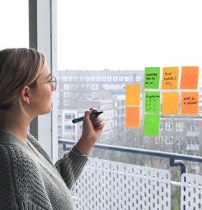 There are a few orange and green post-its notes on the transparent all. A woman is holding on to a pen and is trying to write something on the post-its notes